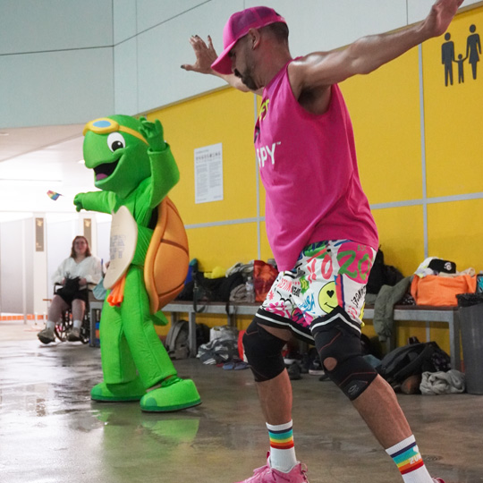 Group fitness instructor wearing a pink outfit an rainbow socks holding a pose with Terry the Turtle the Aquatics Mascot mirroring the pose in the background